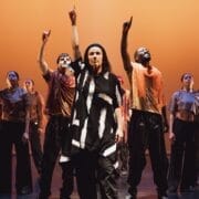 A group of dancers stands on stage under warm orange lighting, embodying the evolution of movement. At the centre, one wears black and white, while others don loose black trousers and sheer orange scarves—many with an arm raised, pointing up. Three60 World energy fills the scene.