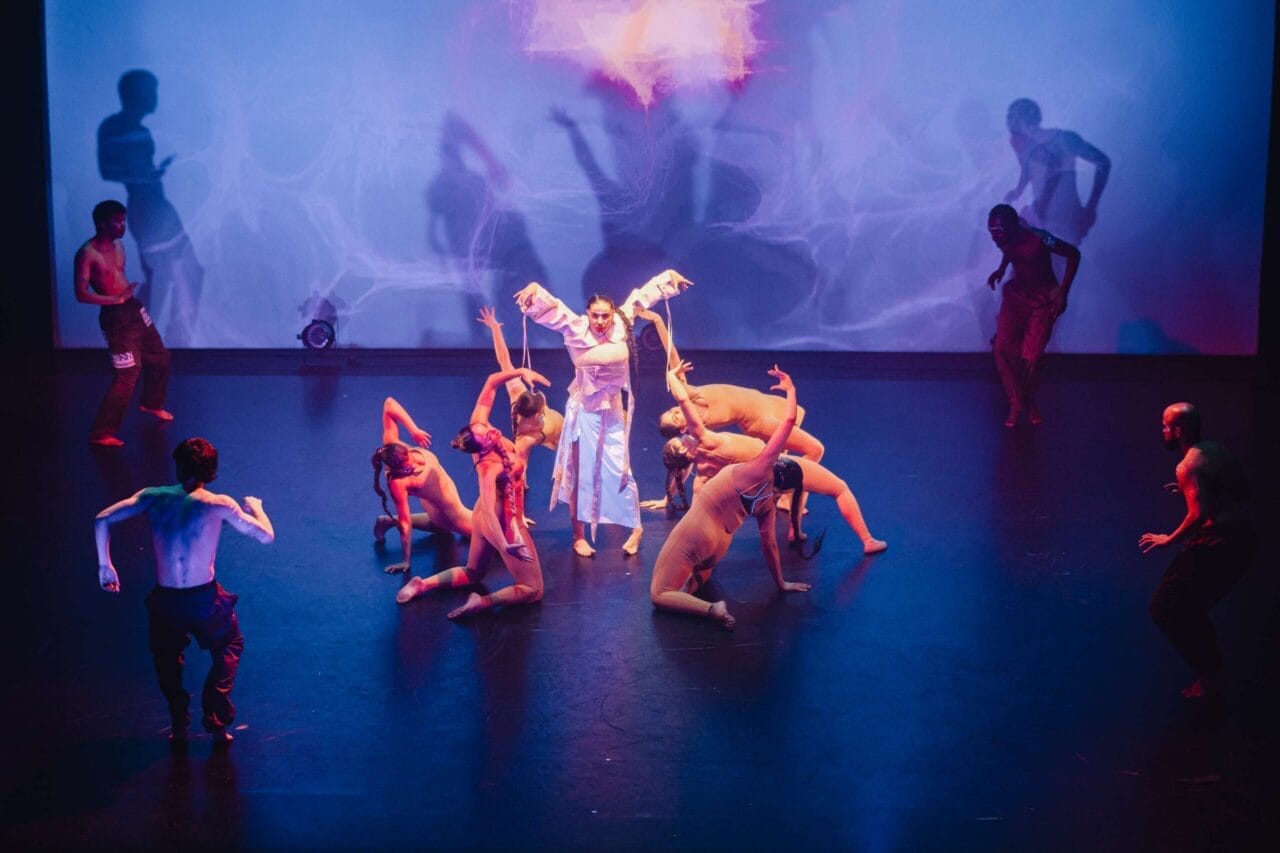 A group of dancers perform on stage under blue and purple lighting in a Three60 World showcase. One person in white stands in the centre with arms raised, embodying evolution, whilst dynamic poses and abstract light patterns fill the background.