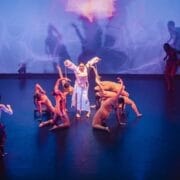 A group of dancers perform on stage under blue and purple lighting in a Three60 World showcase. One person in white stands in the centre with arms raised, embodying evolution, whilst dynamic poses and abstract light patterns fill the background.
