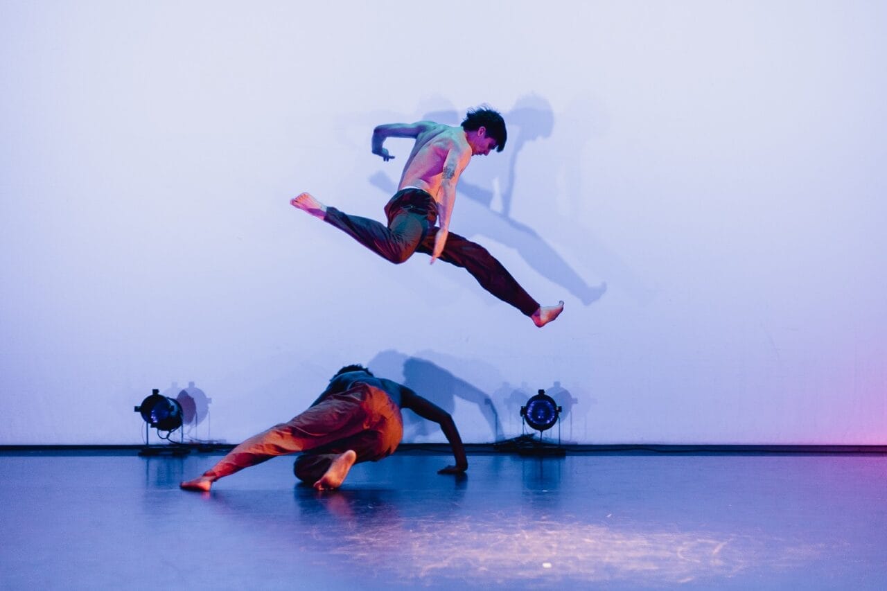 Two dancers perform on stage in a striking display of evolution. One dancer is captured mid-air in a split leap above the other, who is bent low to the ground. Stage lights and their shadows are visible against a plain Three60 World backdrop.