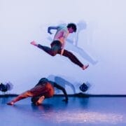 Two dancers perform on stage in a striking display of evolution. One dancer is captured mid-air in a split leap above the other, who is bent low to the ground. Stage lights and their shadows are visible against a plain Three60 World backdrop.