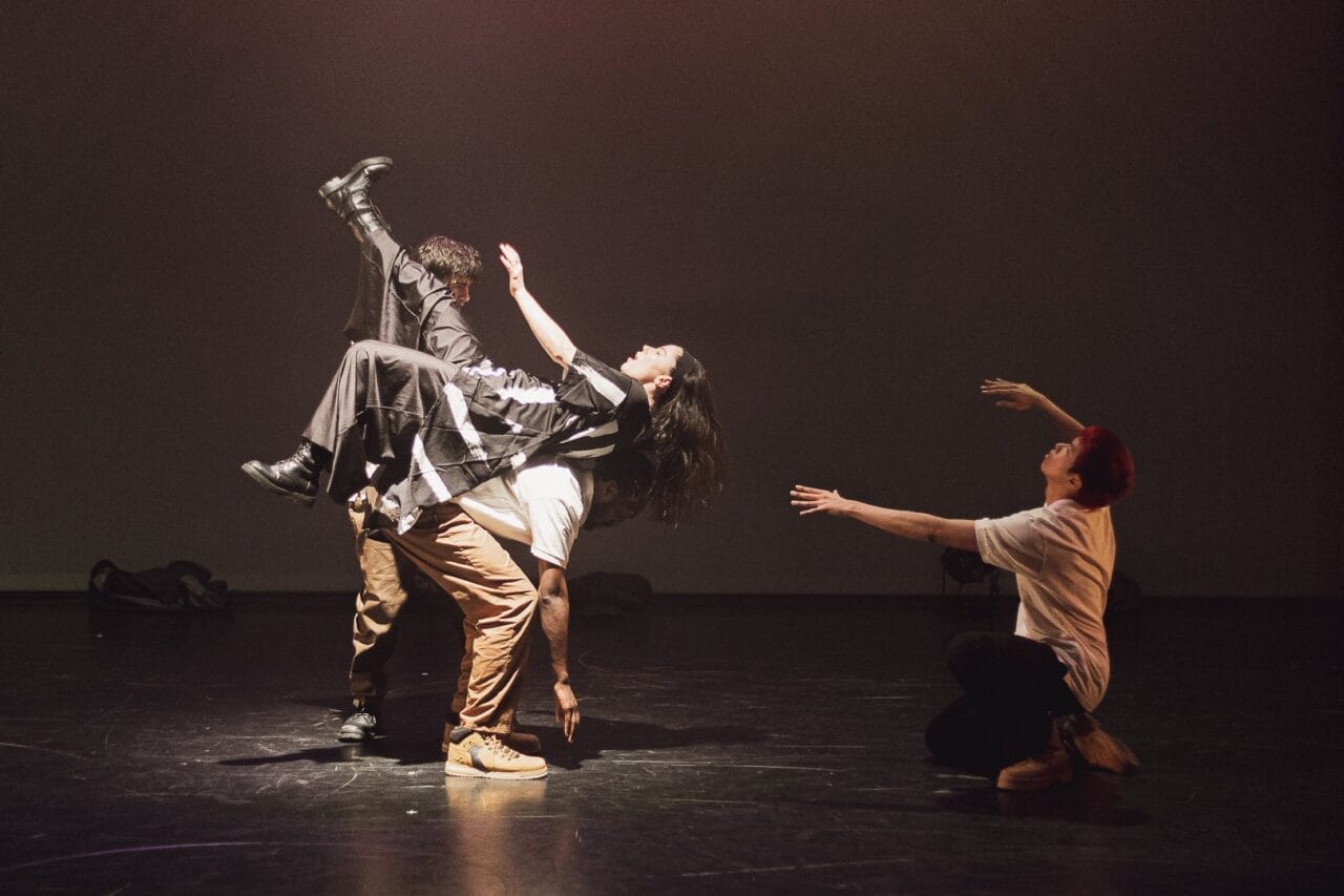 Four dancers perform on stage in a captivating display of Evolution. Two stand and lift a third horizontally in mid-air, whilst a fourth kneels nearby with arms extended upward. The Three60 World stage is dimly lit with a plain background.
