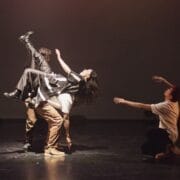 Four dancers perform on stage in a captivating display of Evolution. Two stand and lift a third horizontally in mid-air, whilst a fourth kneels nearby with arms extended upward. The Three60 World stage is dimly lit with a plain background.