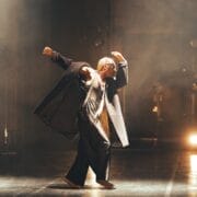 A person with long blond plaits, wearing a black coat and trousers, barefoot, poses with arms raised and bent on a dimly lit stage with spotlights in the background—capturing the spirit of Evolution at Three60 World.