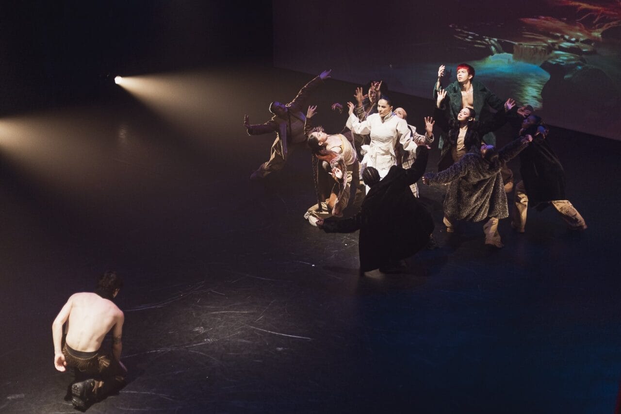 A group of performers in dramatic costumes gather in a spotlight onstage, reaching their arms upwards, whilst a shirtless person kneels alone on the dark floor, facing the group—capturing a striking moment of Evolution by Three60 World.