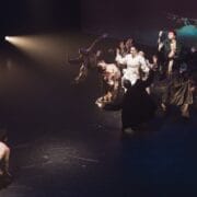 A group of performers in dramatic costumes gather in a spotlight onstage, reaching their arms upwards, whilst a shirtless person kneels alone on the dark floor, facing the group—capturing a striking moment of Evolution by Three60 World.