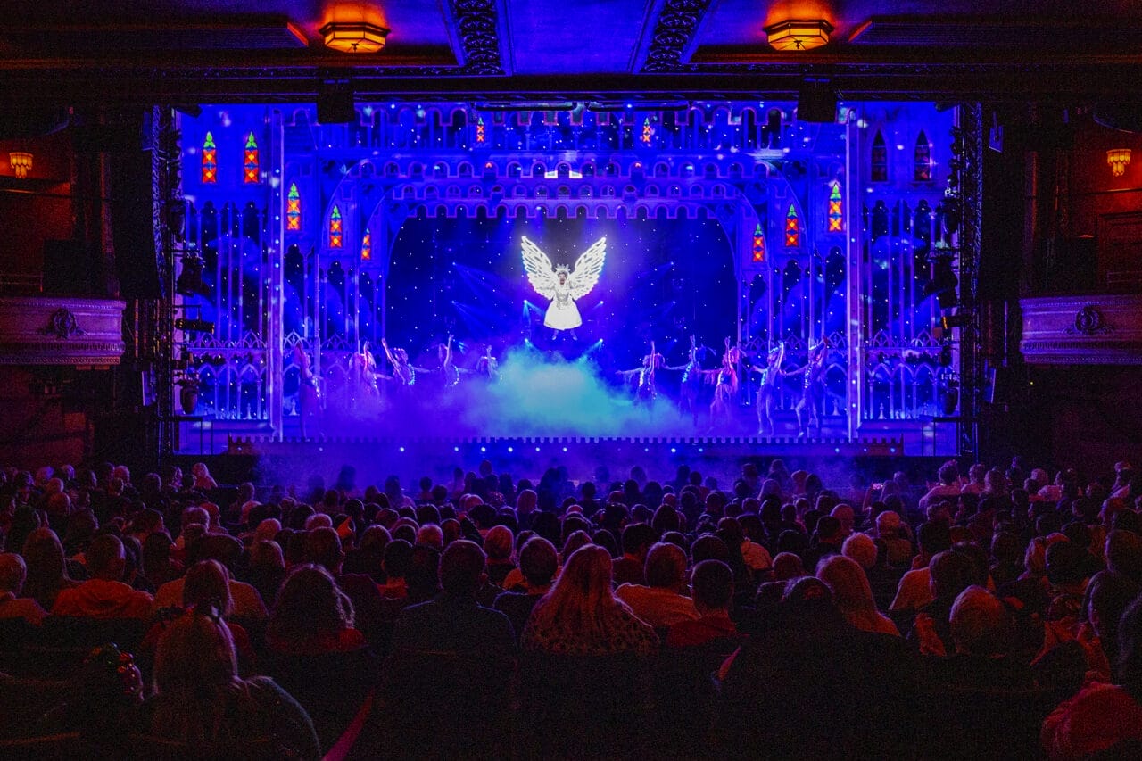 A theater audience watches a stage performance featuring a glowing angel figure and dancers in a brightly lit panto set with blue and purple tones, celebrating 50 years of magic starring Allan Stewart.