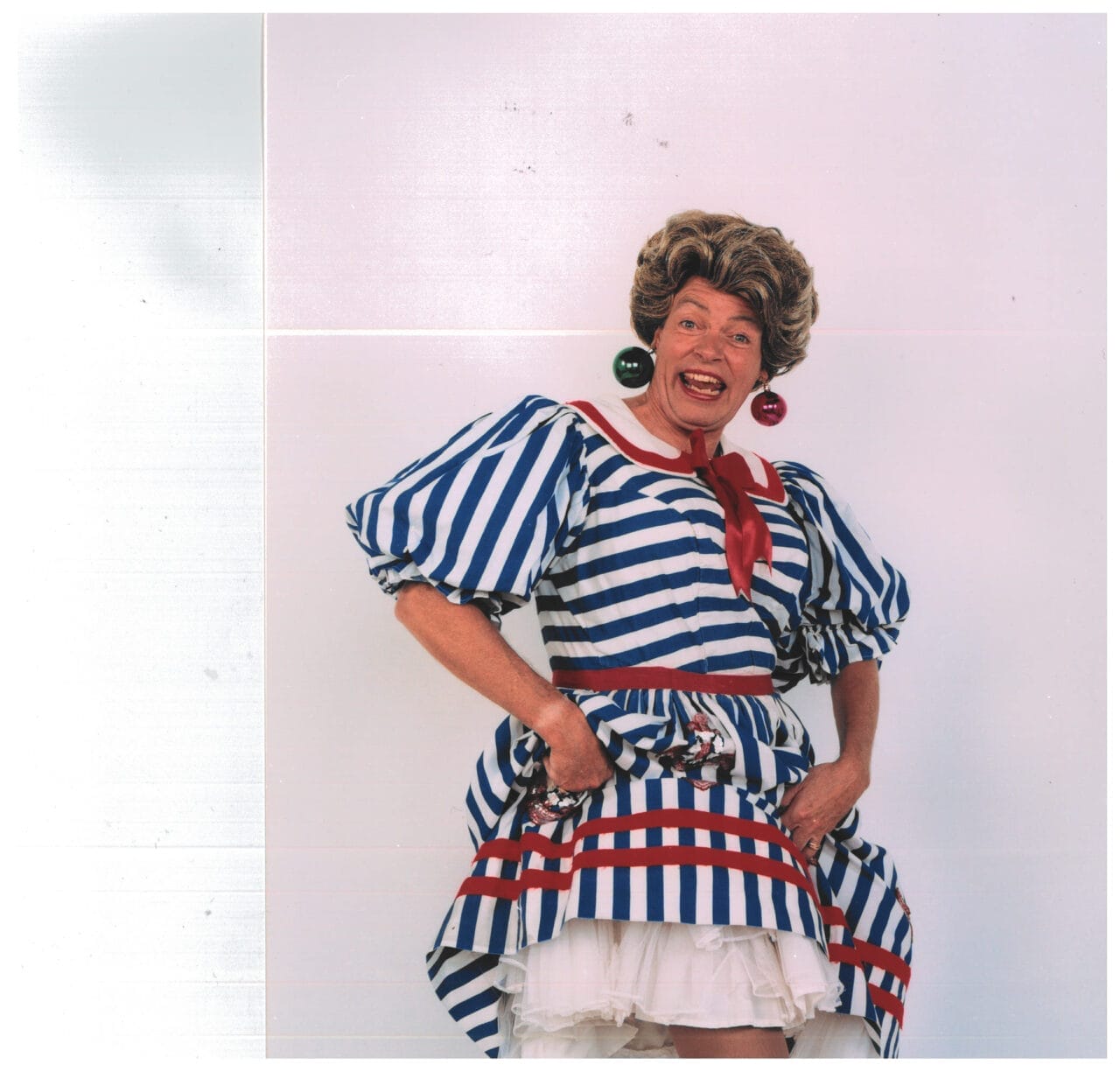 A person dressed in a blue and white striped dress with puffed sleeves, a red scarf, and large red and green earrings stands smiling in front of a plain background, celebrating 50 years of panto like Allan Stewart.
