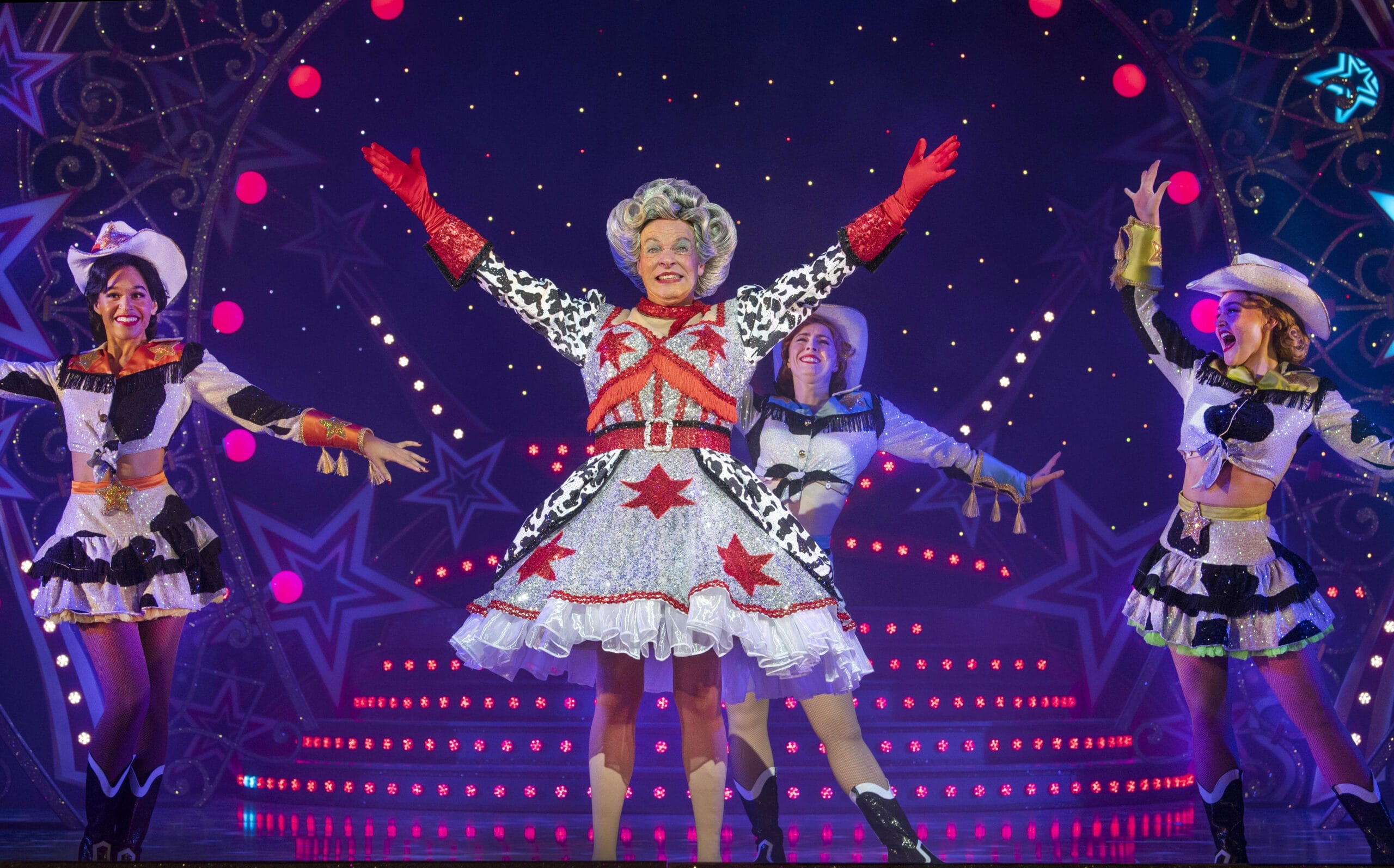 Four performers in colorful, star-themed costumes celebrate 50 years of panto as they pose on a brightly lit stage with Allan Stewart, surrounded by dazzling lights and star decorations.