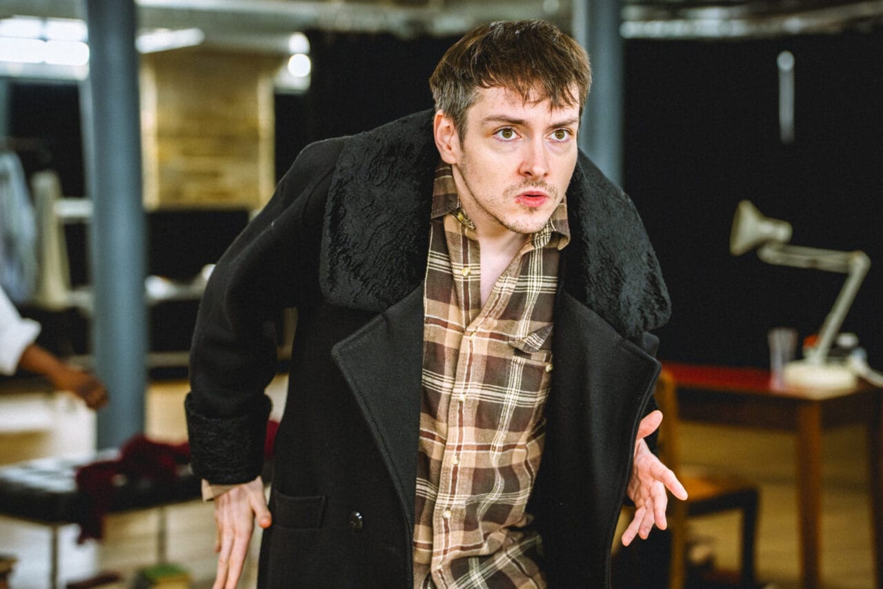 A man with short brown hair and a checked shirt under a dark coat leans forward with a focused expression, as if contemplating crime and punishment, in an indoor setting with tables and chairs in the background.