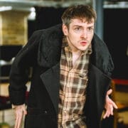 A man with short brown hair and a checked shirt under a dark coat leans forward with a focused expression, as if contemplating crime and punishment, in an indoor setting with tables and chairs in the background.