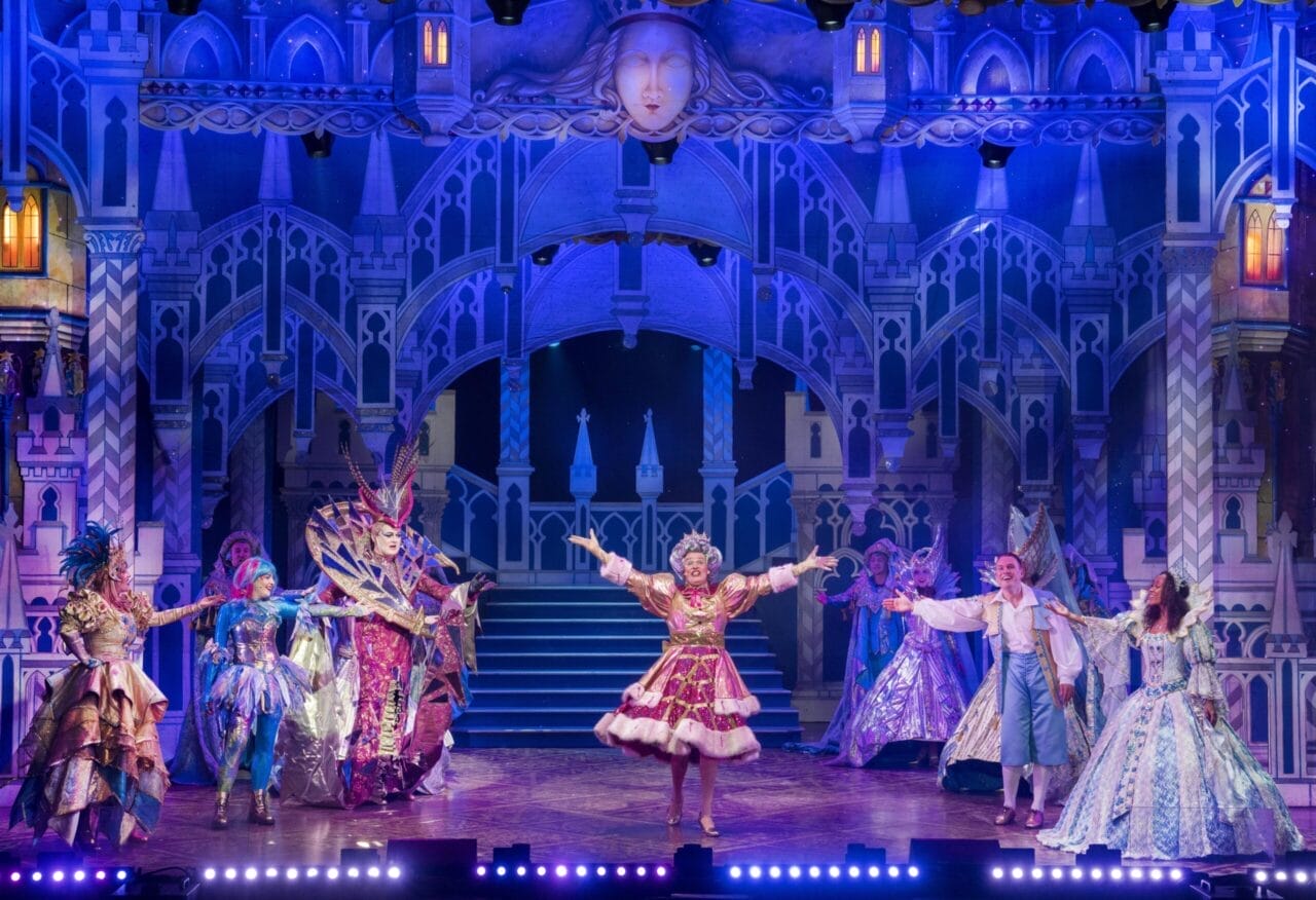A group of performers in elaborate, colorful costumes act on a brightly lit stage with a castle-themed backdrop, celebrating 50 years of panto magic with Allan Stewart.