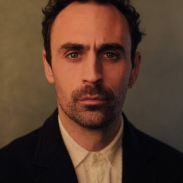 A man with short dark hair, a trimmed beard, and an earring wears a dark coat over a buttoned white shirt, looking directly at the camera with a neutral expression