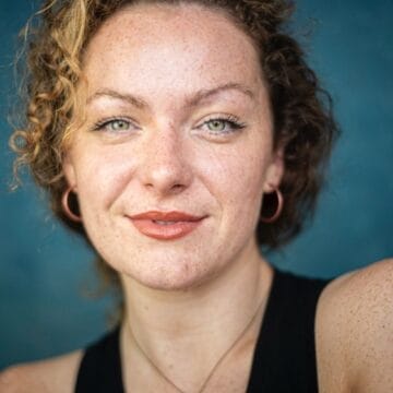 A woman with curly hair, hoop earrings, and a black top is smiling slightly at the camera in front of a blue background