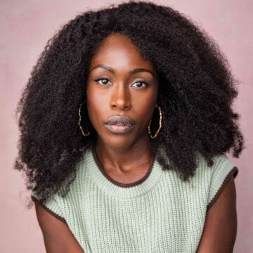 Woman with natural curly hair and hoop earrings wearing a sleeveless knit top, looking directly at the camera against a pale pink background