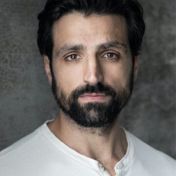 A man with dark hair and a full beard, wearing a white henley shirt, looks at the camera with a neutral expression against a gray textured background, reminiscent of a scene from The Constant Wife starring Kara Tointon.