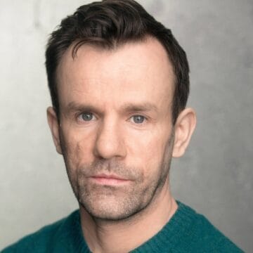 A man with short brown hair and light stubble, dressed in a teal sweater, looks directly at the camera against a neutral background