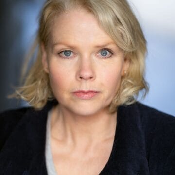 A woman with short blonde hair and blue eyes looks directly at the camera, wearing a dark jacket over a light top.