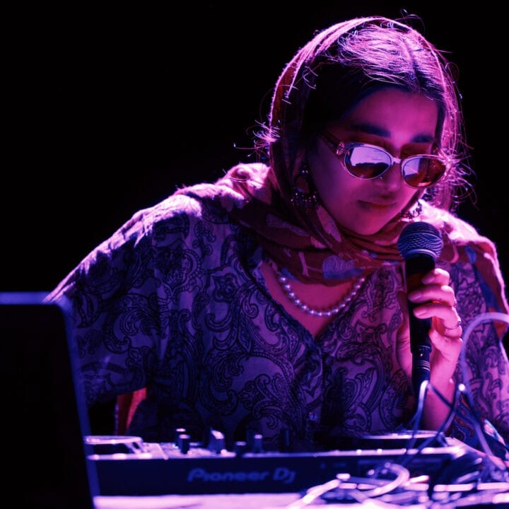 A person wearing sunglasses and a patterned headscarf holds a microphone while operating DJ equipment under purple lighting.
