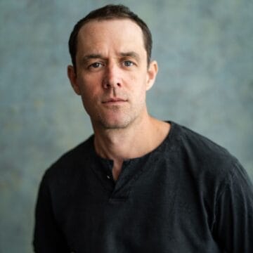 A man with short brown hair, wearing a dark t-shirt, looks directly at the camera against a soft, mottled background