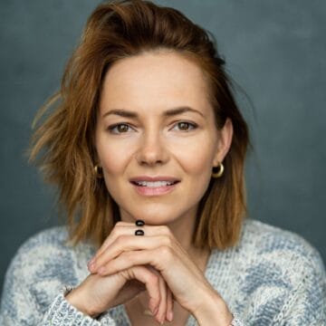 A woman with short, light brown hair and gold hoop earrings rests her chin on her hands, wearing a gray knit sweater and smiling slightly in front of a plain background
