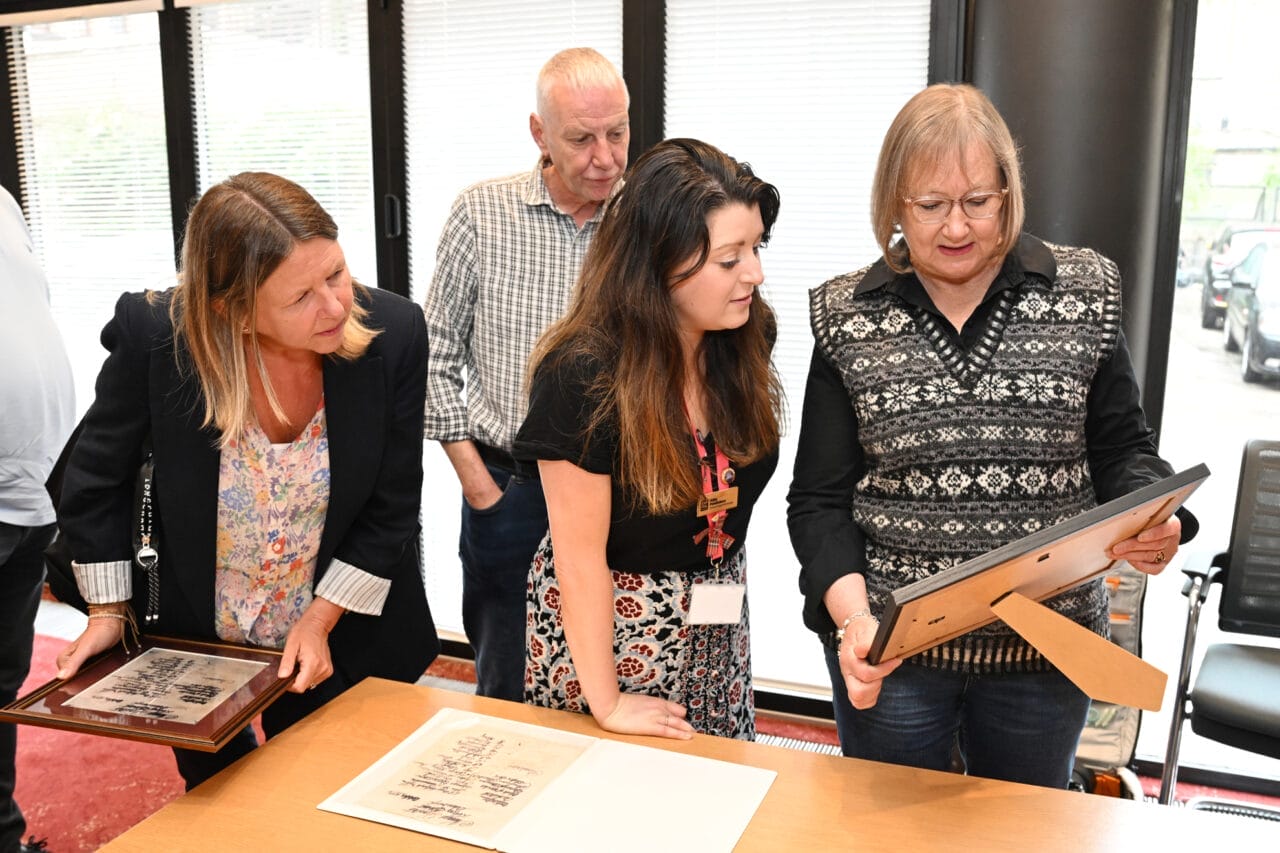 Four adults stand together indoors at a Volunteer Event, examining framed photographs or documents on a table; one woman points at an item while others observe.