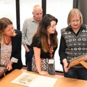 Four adults stand together indoors at a Volunteer Event, examining framed photographs or documents on a table; one woman points at an item while others observe.