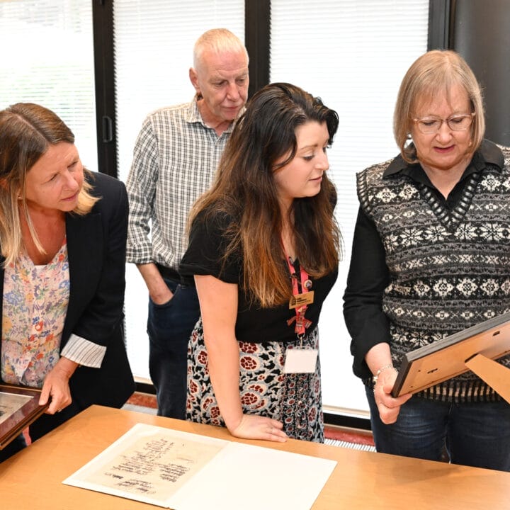 Four adults stand together indoors at a Volunteer Event, examining framed photographs or documents on a table; one woman points at an item while others observe.