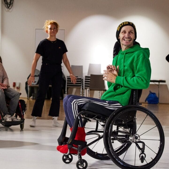 A group of five people, two in wheelchairs and three standing, are gathered in a well-lit room with chairs in the background.