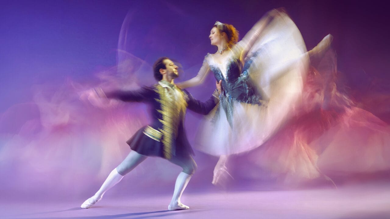 A male and a female ballet dancer perform on stage. The man supports the woman as she leaps mid-air, her tutu and arms creating a motion blur. The background is purple with additional blurred dancers visible.