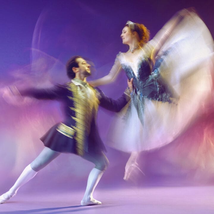 A male and a female ballet dancer perform on stage. The man supports the woman as she leaps mid-air, her tutu and arms creating a motion blur. The background is purple with additional blurred dancers visible.
