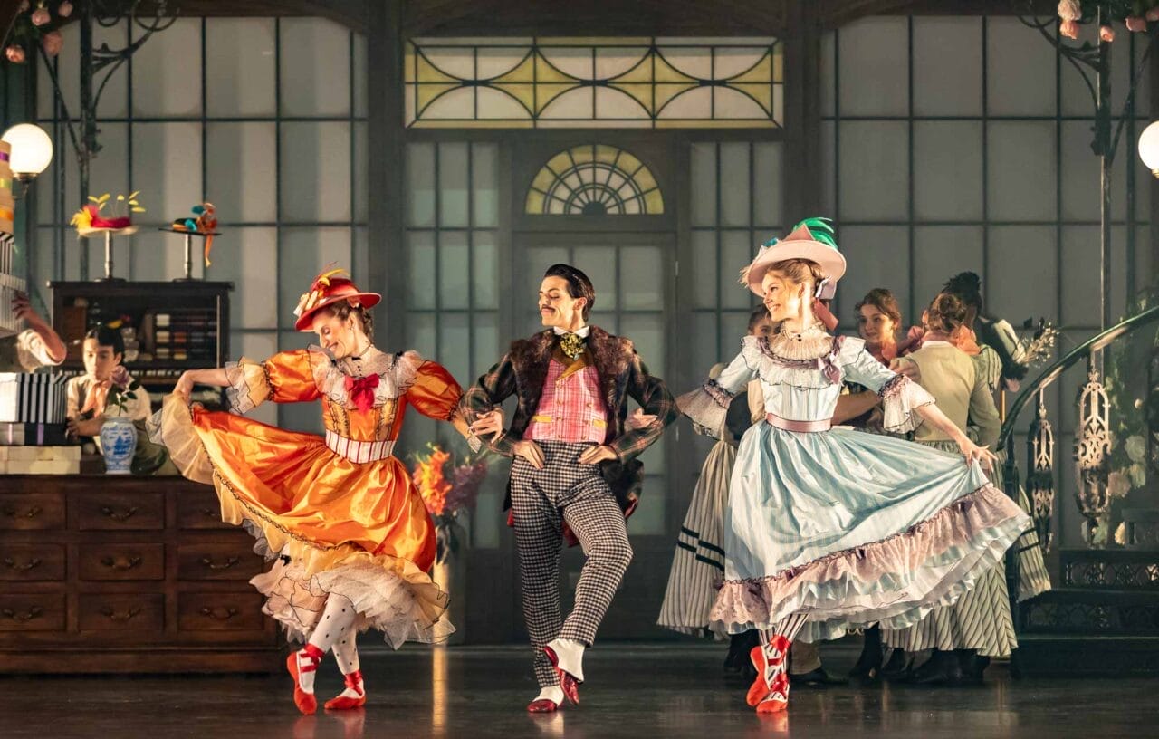 Three performers in colourful period costumes dance on stage; two women wear bright dresses and hats, while a man in a suit and bow tie dances between them.