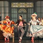 Three performers in colourful period costumes dance on stage; two women wear bright dresses and hats, while a man in a suit and bow tie dances between them.