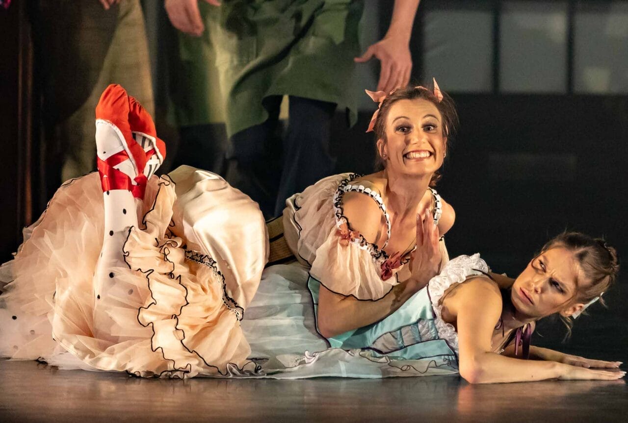 Two women in colourful, ruffled costumes lie on the stage floor during a performance. The woman on top smiles widely with hands clasped, while the woman below looks to the side with a displeased expression.