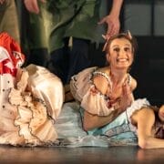Two women in colourful, ruffled costumes lie on the stage floor during a performance. The woman on top smiles widely with hands clasped, while the woman below looks to the side with a displeased expression.