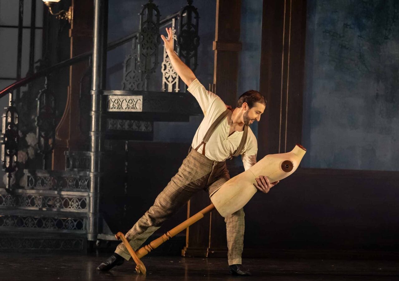 A man in period clothing leans back while holding a wooden dress form, performing on stage