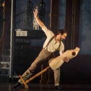 A man in period clothing leans back while holding a wooden dress form, performing on stage