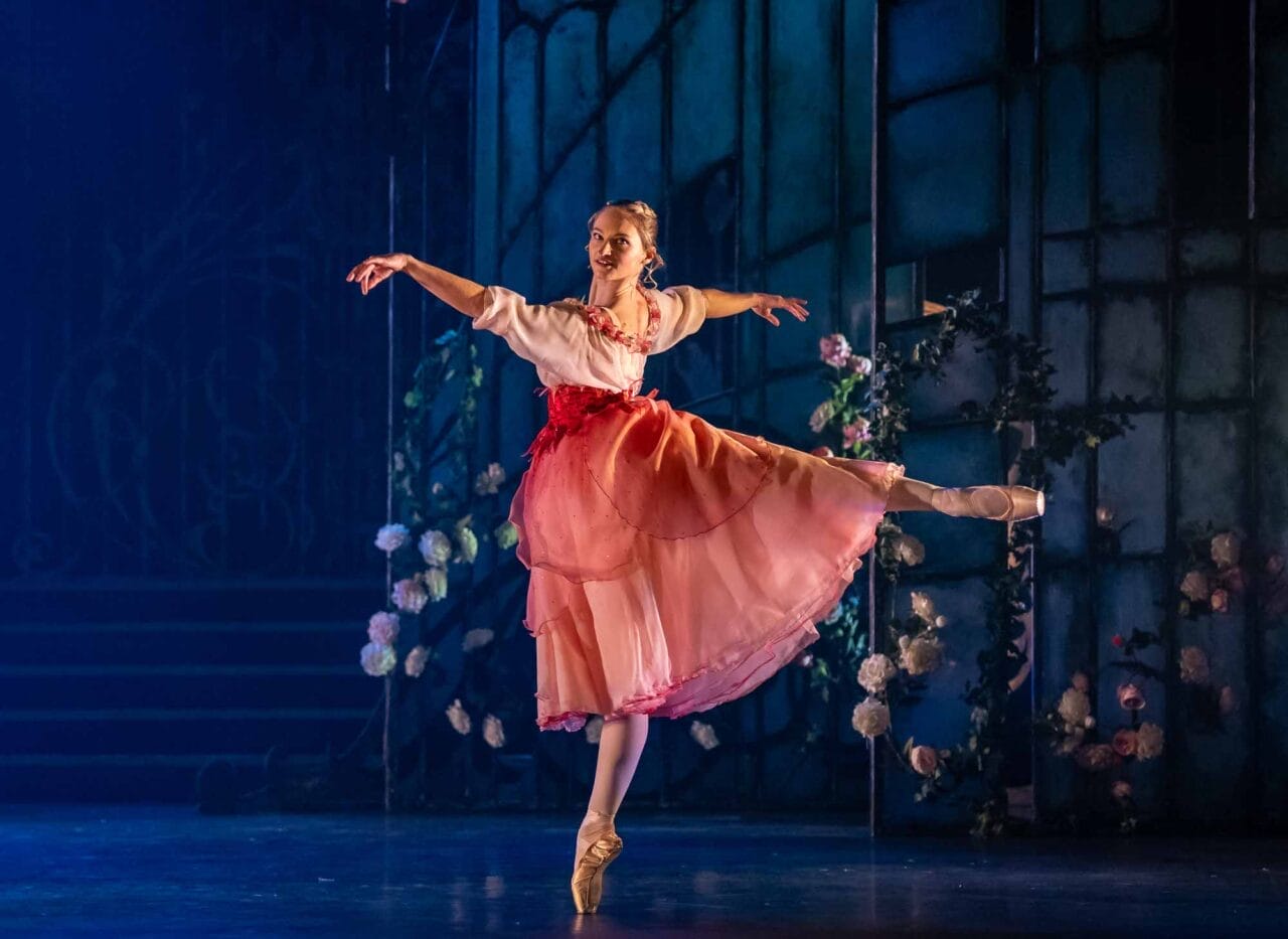A ballerina wearing a pink and red costume stands en pointe with one leg extended behind her and arms gracefully outstretched on a stage decorated with flowers and backdrop panels.