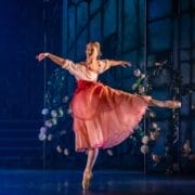 A ballerina wearing a pink and red costume stands en pointe with one leg extended behind her and arms gracefully outstretched on a stage decorated with flowers and backdrop panels.