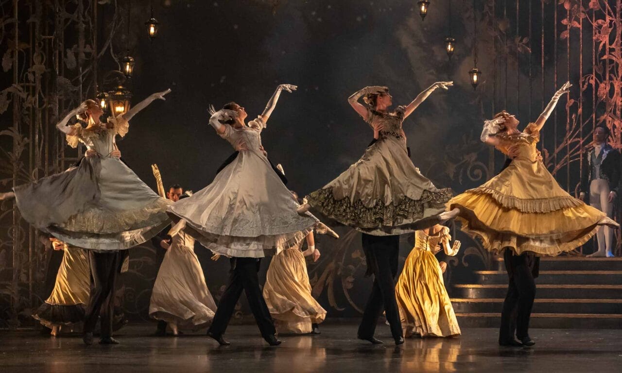 A group of dancers in period costumes with long flowing skirts perform a synchronised ballet move on stage, with their arms raised and skirts lifted.