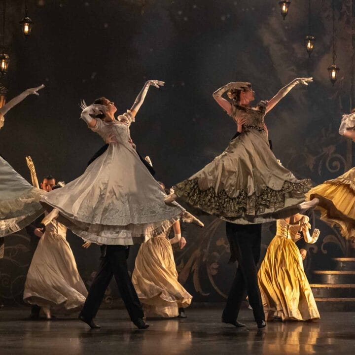 A group of dancers in period costumes with long flowing skirts perform a synchronised ballet move on stage, with their arms raised and skirts lifted.