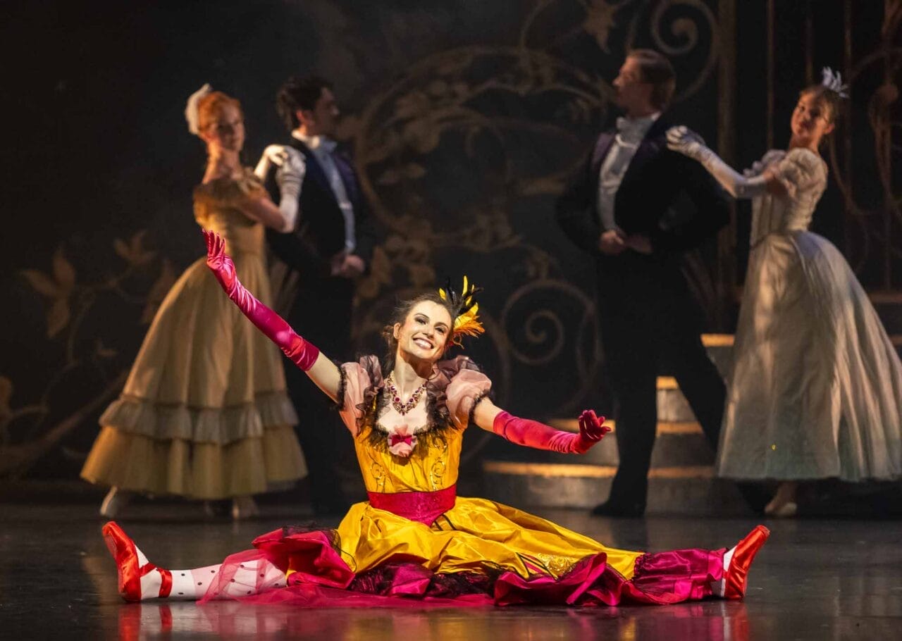 A dancer in a colourful yellow and pink costume sits in a split with arms outstretched, smiling.