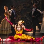 A dancer in a colourful yellow and pink costume sits in a split with arms outstretched, smiling.