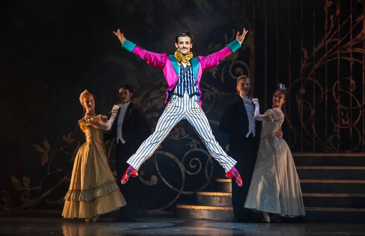 A performer in a bright pink jacket, striped trousers, and bow tie jumps with arms and legs outstretched on stage