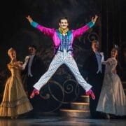 A performer in a bright pink jacket, striped trousers, and bow tie jumps with arms and legs outstretched on stage