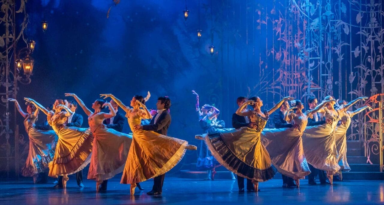 A group of dancers in formal costumes perform a synchronised dance on stage, with women in flowing dresses and men in suits.