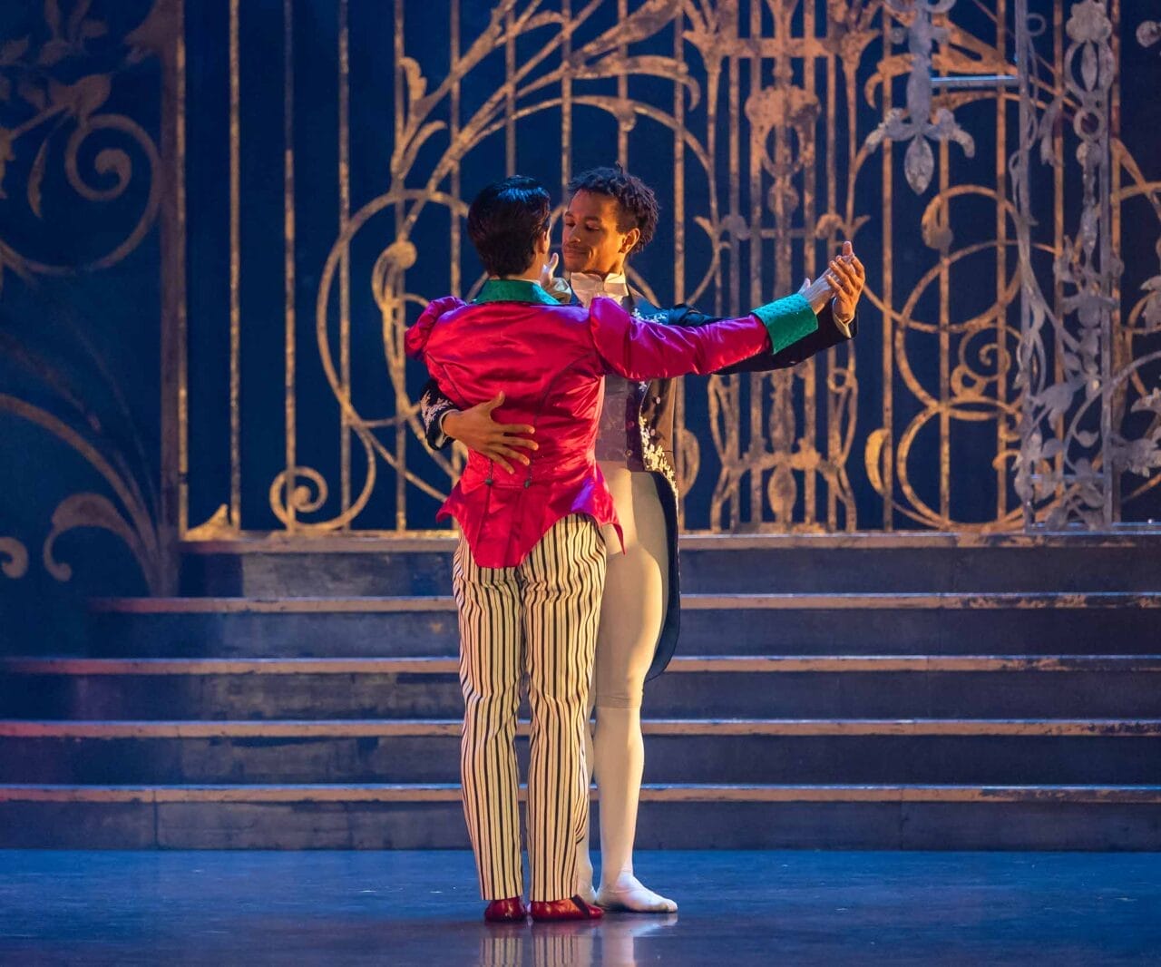 Two male ballet dancers perform a duet on stage, with one wearing a bright pink jacket and striped trousers and the other in a purple jacket and white tights.