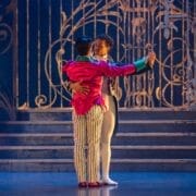 Two male ballet dancers perform a duet on stage, with one wearing a bright pink jacket and striped trousers and the other in a purple jacket and white tights.