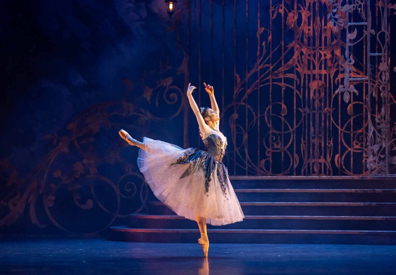 A ballerina in a sparkling tutu performs a ballet pose on stage, standing on one pointed foot with the other leg extended behind her.