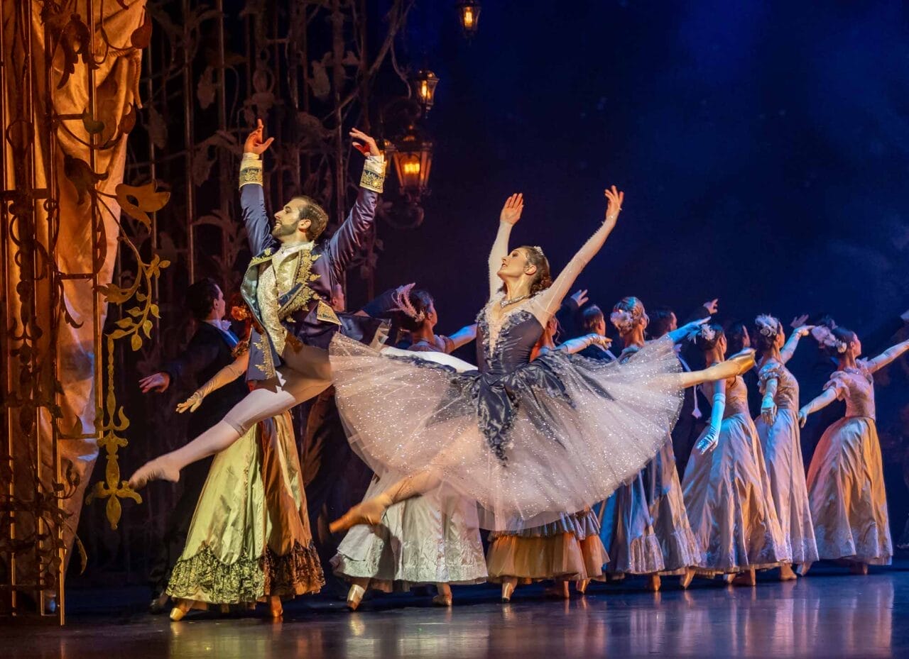 Ballet dancers perform on stage in elaborate costumes, with two lead dancers leaping gracefully at the front